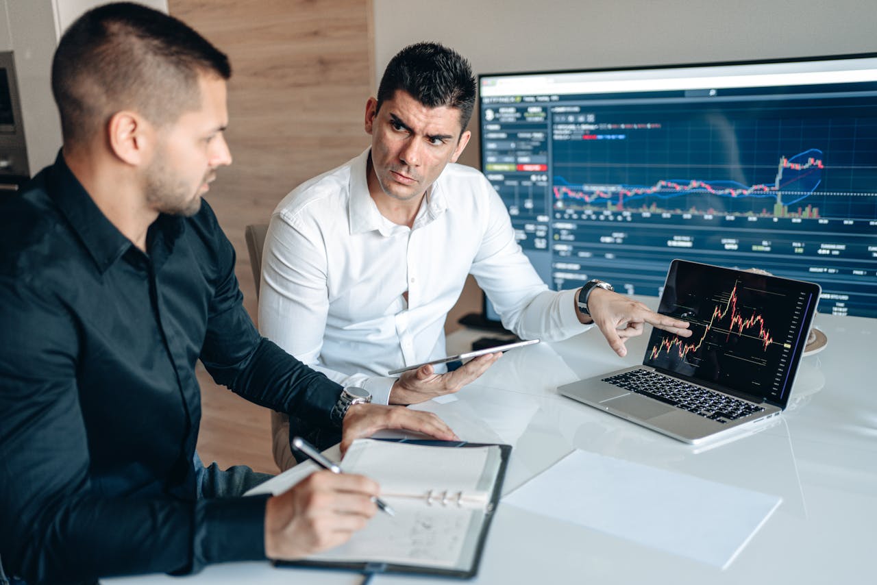 Two businessmen discuss financial charts on laptops in an office setting.